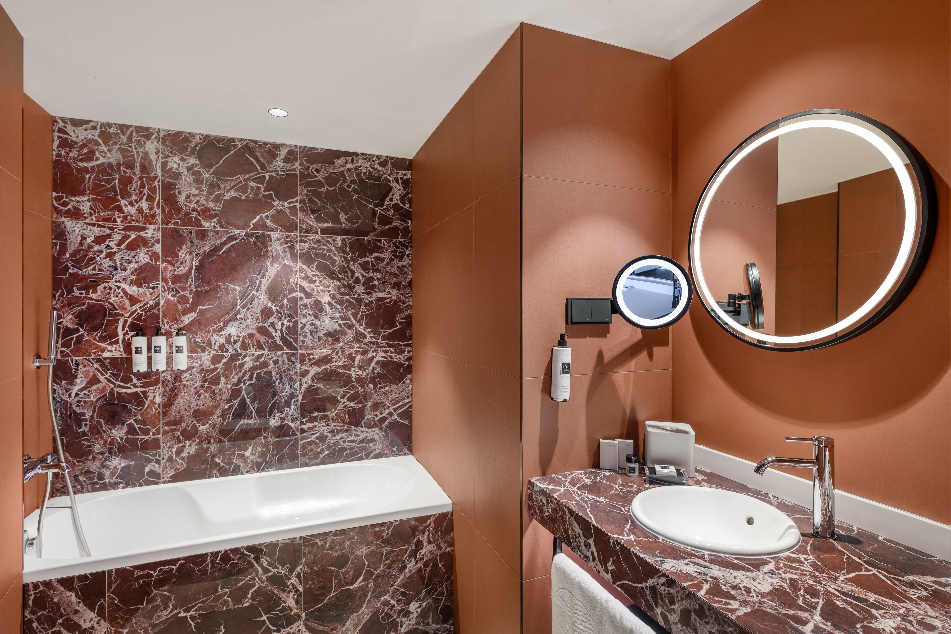 A modern bathroom with a marble wall, a white bathtub, a sink with a mirror, and toiletries on the wall.