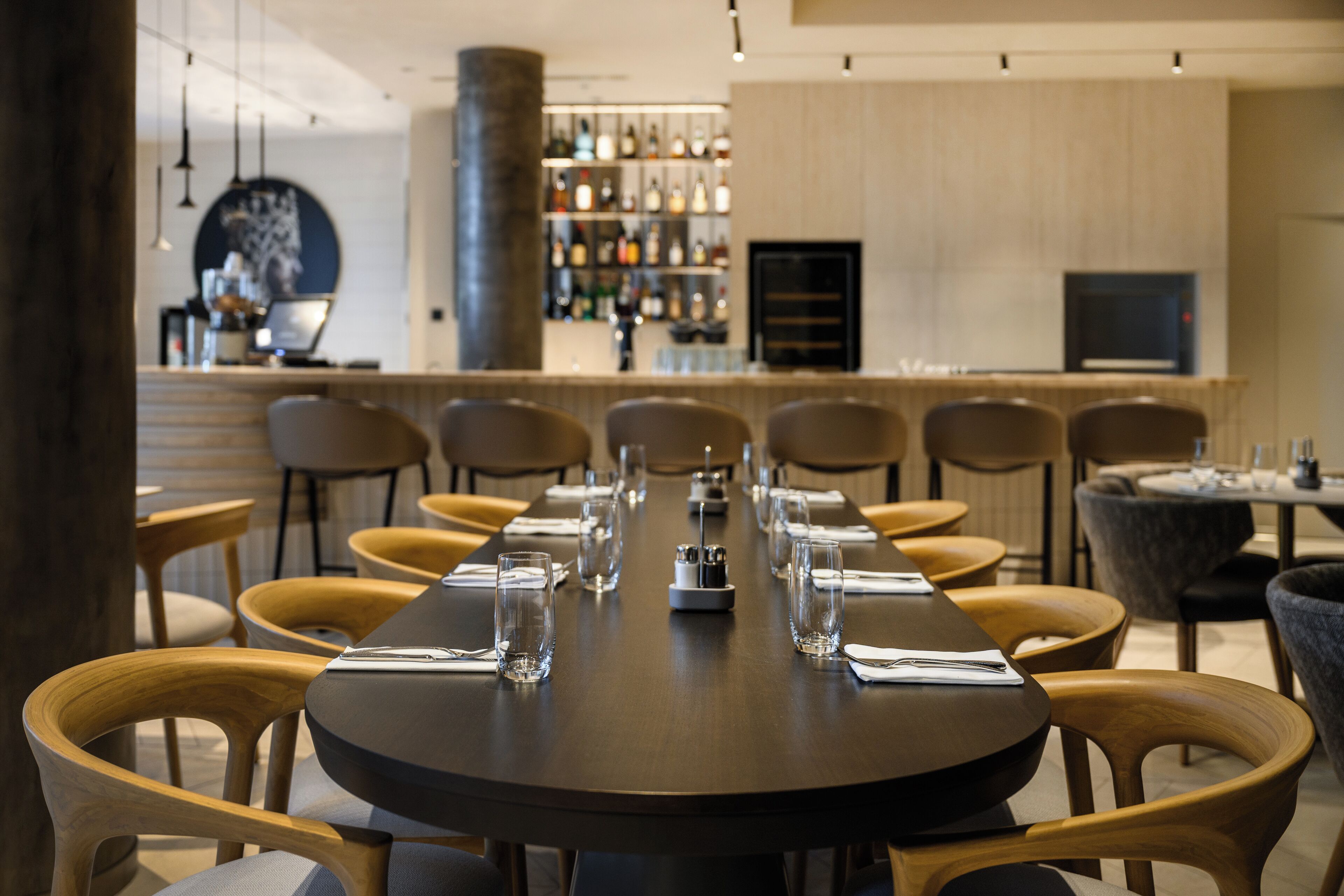 A restaurant with a long table set for a meal, wooden chairs, a bar area in the background, and a modern interior design.