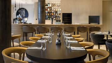 A restaurant with a long table set for a meal, wooden chairs, a bar area in the background, and a modern interior design.