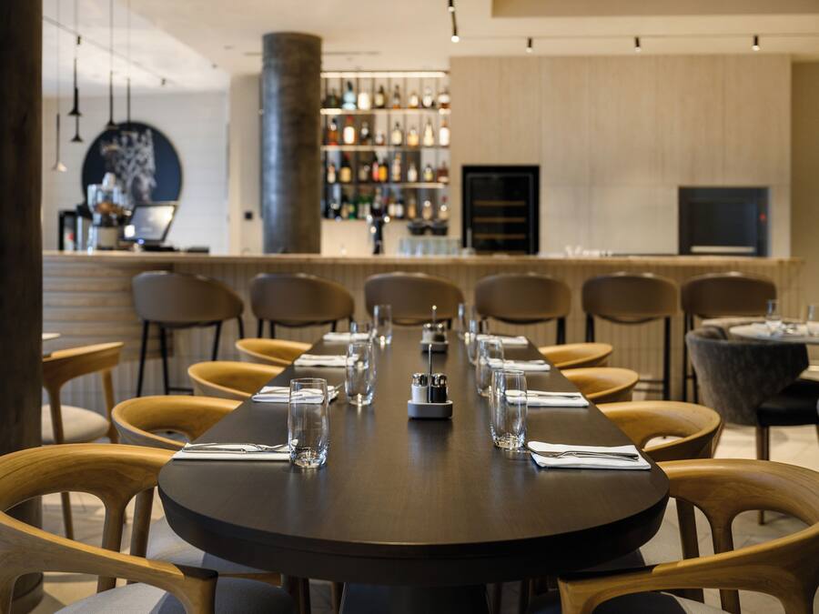 Ein Restaurant mit einem langen Tisch, gedeckt für eine Mahlzeit, Holzsesseln, einem Barbereich im Hintergrund und einem modernen Innenraum.