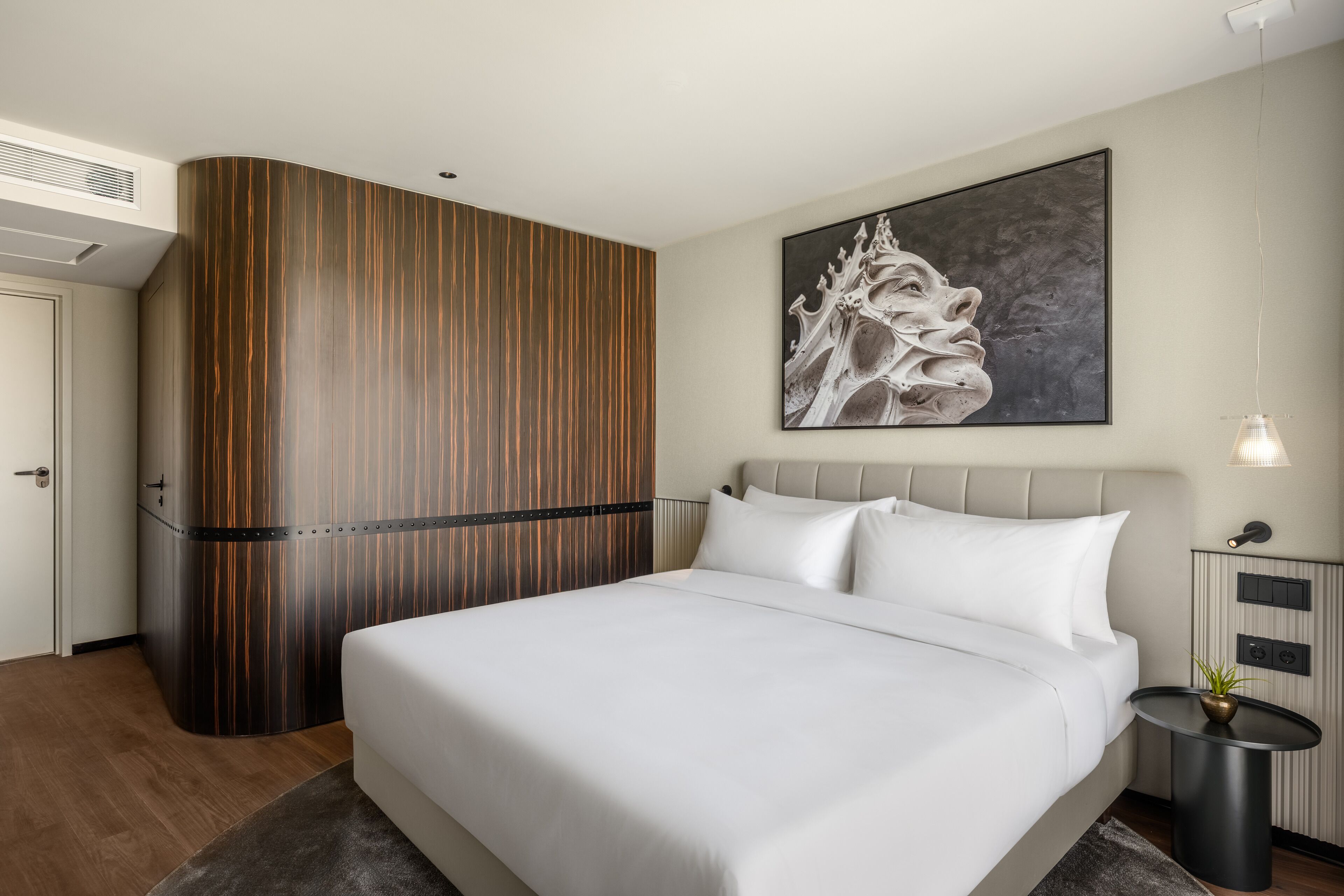 A modern hotel room with a large bed, a wooden accent wall, a painting, a bedside table with a lamp, and a small plant.