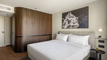 A modern hotel room with a large bed, a wooden accent wall, a painting, a bedside table with a lamp, and a small plant.