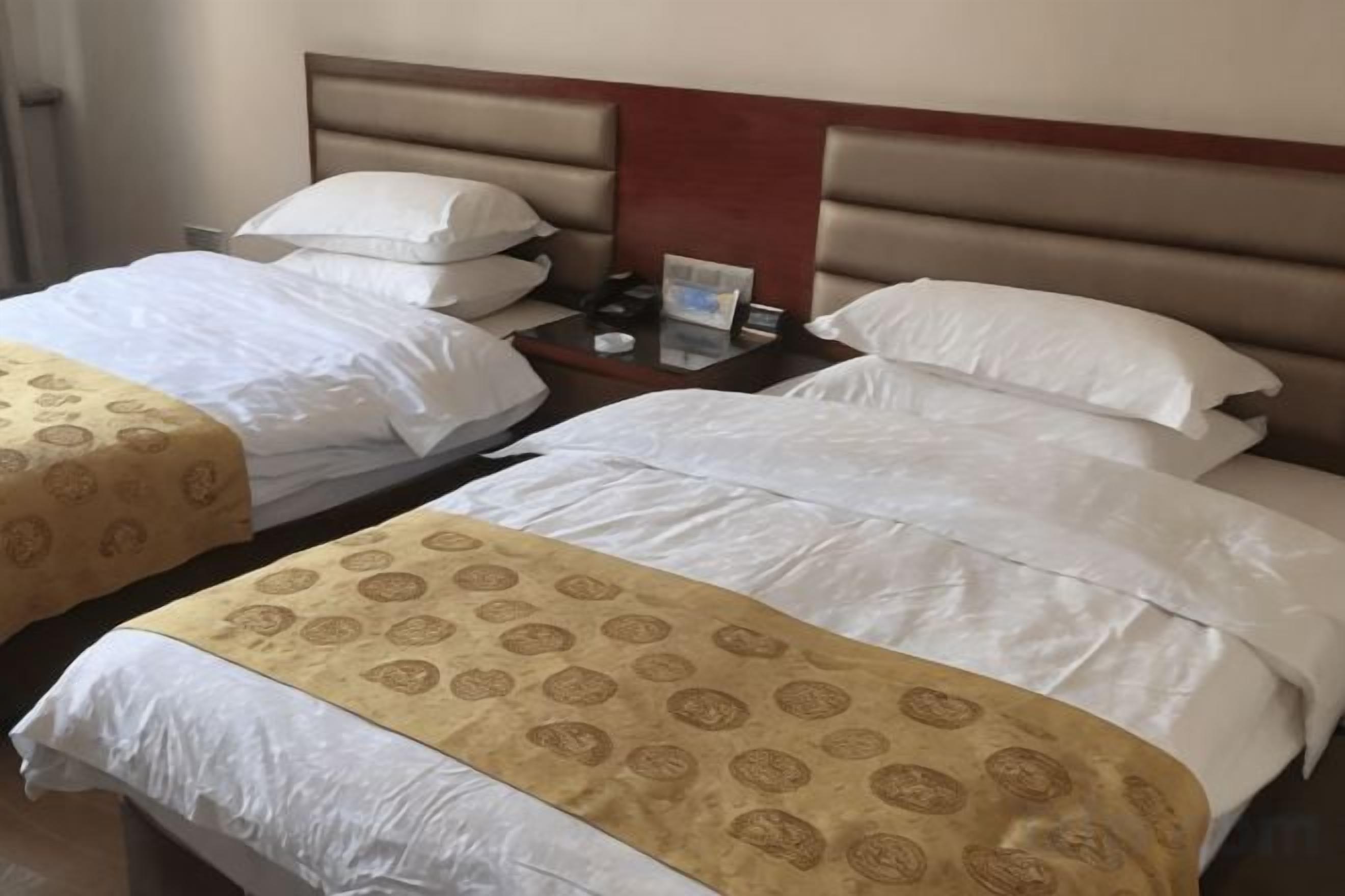 A hotel room with two beds, each with a headboard and pillows. There is a nightstand with a clock and a lamp between the beds.