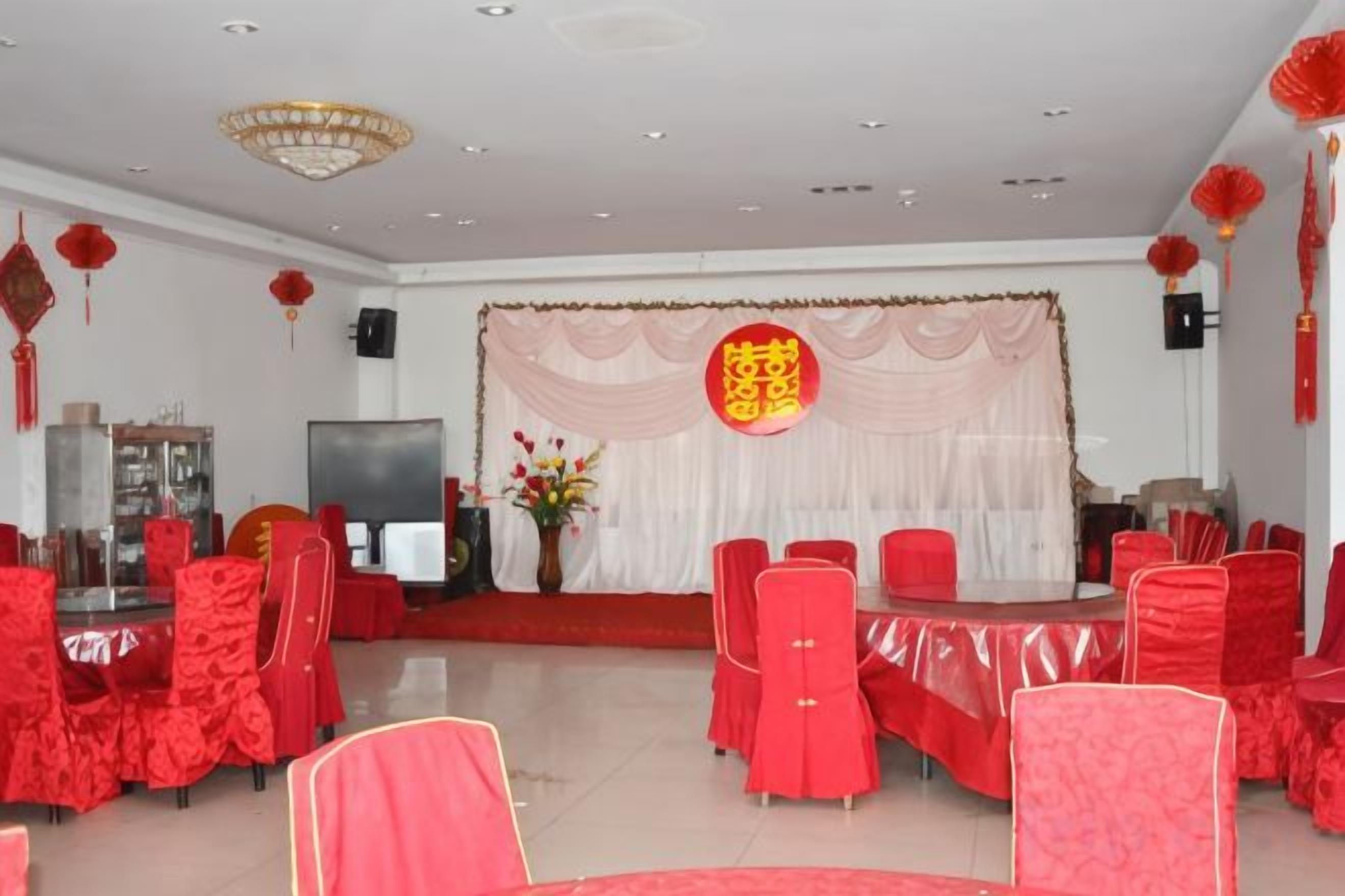 Un salón de banquetes con mesas y sillas redondas, un escenario con un telón de fondo y decoraciones rojas.