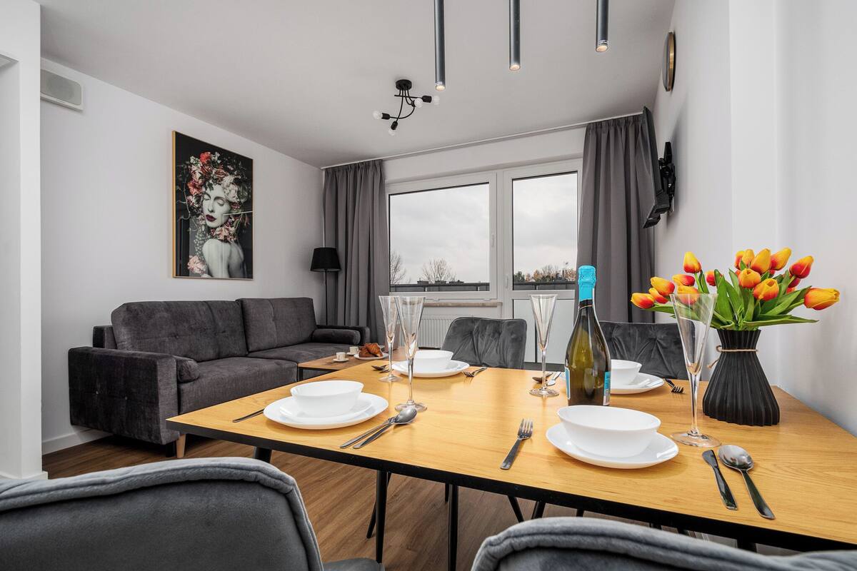 A modern dining area with a wooden table set for four, a gray sofa, and a large window with curtains.