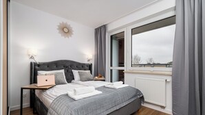 A modern bedroom with a grey bed, a laptop on the nightstand, and a view of the city through the window. - Pileckiego Residence (Warsaw)