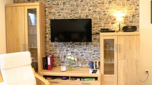 A living room with a stone wall, a flat-screen TV, wooden cabinets, a chair, and a small table with books and a vase. - Ostersander vacation apartment - centrally located between Ihlow, Timmel, Aurich and bathing lakes (Ihlow)