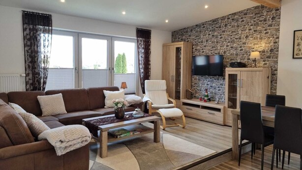 A living room with a brown sofa, a white armchair, a coffee table, and a TV mounted on a stone wall. - Ostersander vacation apartment - centrally located between Ihlow, Timmel, Aurich and bathing lakes (Ihlow)