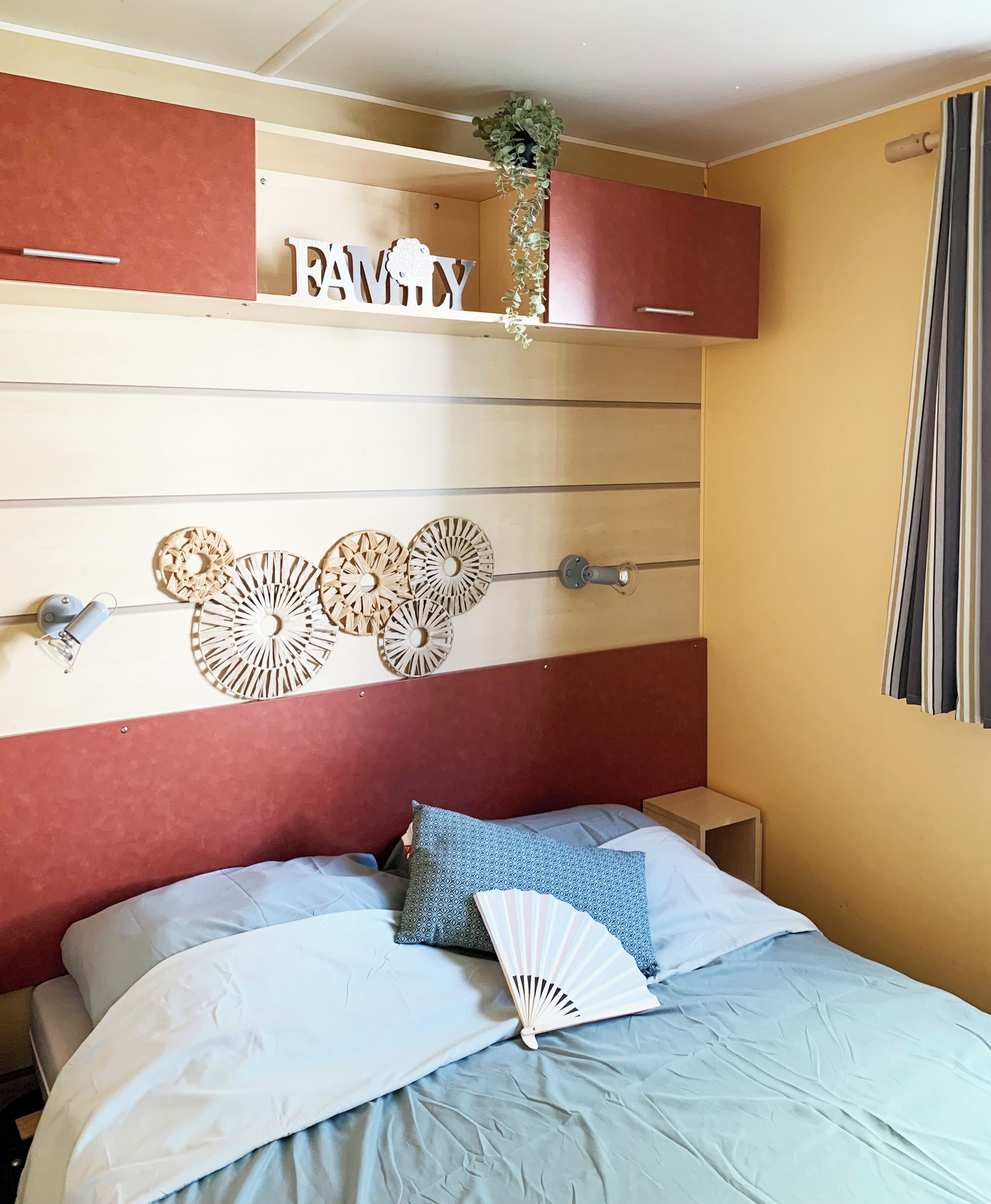 Quarto com cama, almofadas, mesa de cabeceira, prateleira na parede com uma planta e um objeto decorativo.