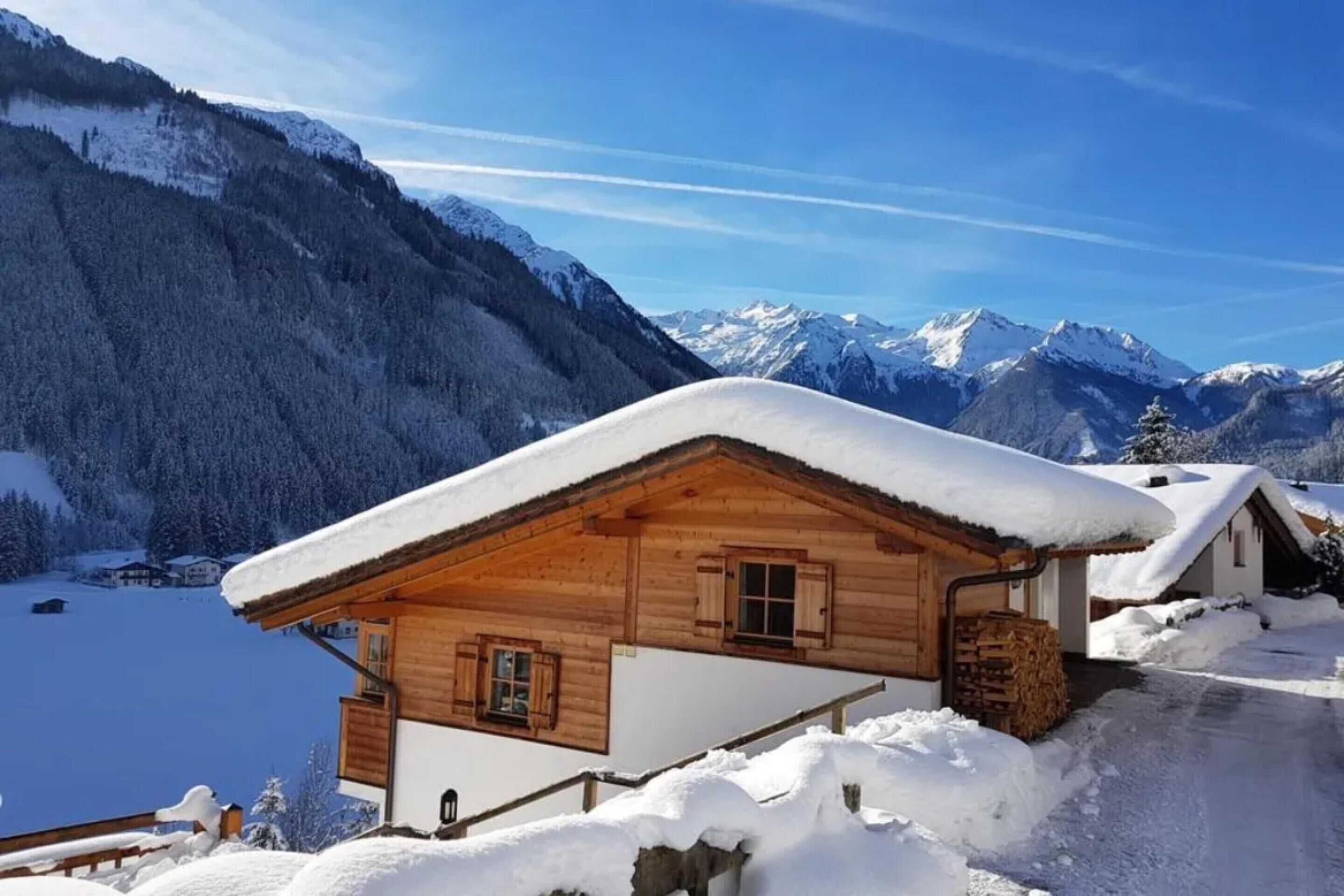 A wooden chalet with a sloped roof covered in snow, surrounded by a snowy landscape and mountains.