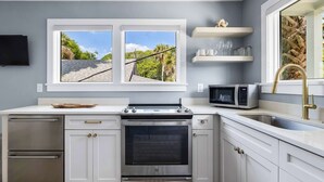 A modern kitchen with white cabinets, a stainless steel oven, a microwave, and a window with a view of trees. - Beautiful Studio with Kitchen + Parking (Folly Beach)