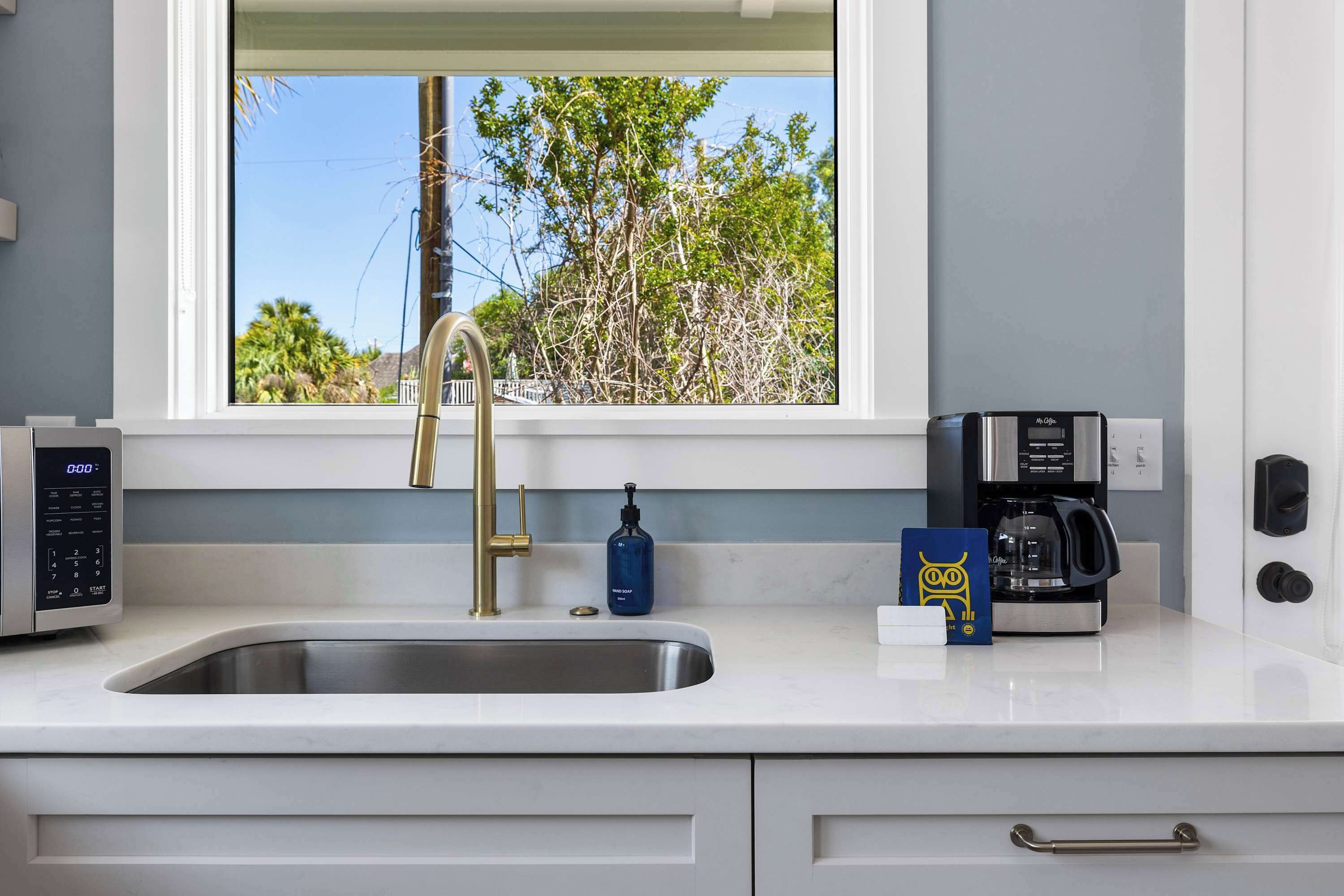 A modern kitchen with a stainless steel sink, a gold faucet, a coffee maker, and a window with a view of trees.