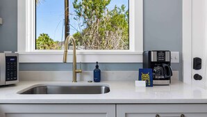 A modern kitchen with a stainless steel sink, a gold faucet, a coffee maker, and a window with a view of trees. - Beautiful Studio with Kitchen + Parking (Folly Beach)