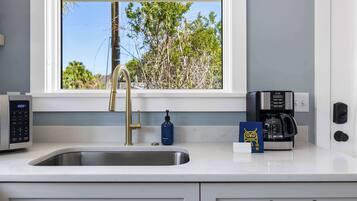 A modern kitchen with a stainless steel sink, a gold faucet, a coffee maker, and a window with a view of trees.