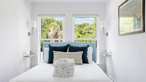 Ein ordentlich gemachtes Bett mit weißen Leinen und einem gefalteten Handtuch, zwei Fenster mit Blick ins Grüne und ein gerahmtes Bild an der Wand.