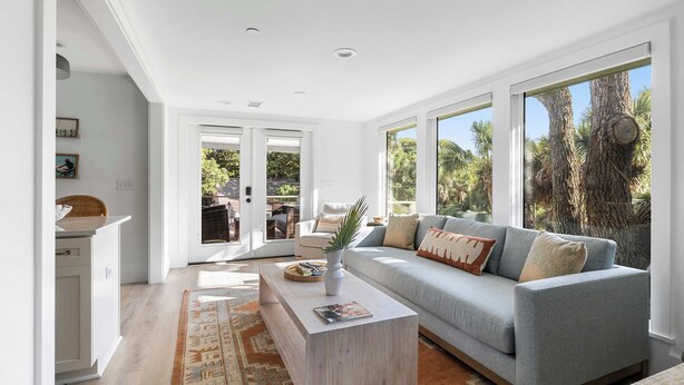 A spacious living room with a grey sofa, a white coffee table, and a large window offering a view of palm trees. - Large, Upgraded 3bd with Parking, steps to beach! (Folly Beach)