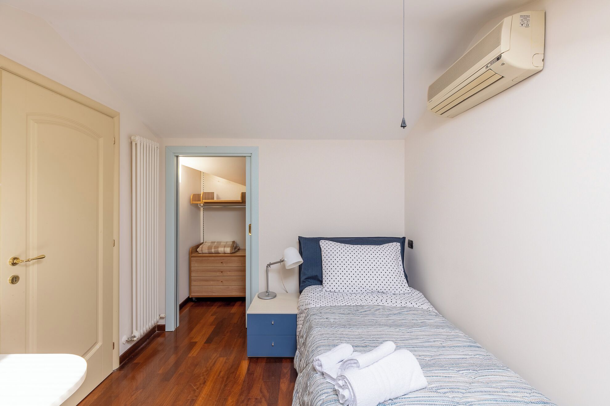 A small, well-lit bedroom with a single bed, a bedside table, a lamp, and a wardrobe.
