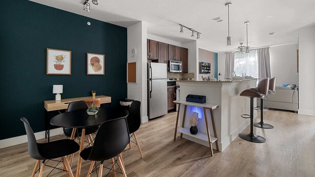 A modern kitchen with wooden cabinets, a refrigerator, and a microwave. There is a dining area with black chairs and a table. - Dallashaus Near Galleria Medical City (Dallas)