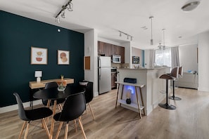 A modern kitchen with wooden cabinets, a refrigerator, and a microwave. There is a dining area with black chairs and a table.