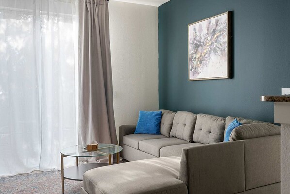 A modern living room with a grey sectional sofa, a glass coffee table, and a blue abstract painting on the wall.