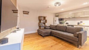 A modern living room with a sectional sofa, a flat-screen TV, wooden flooring, and a kitchen in the background.