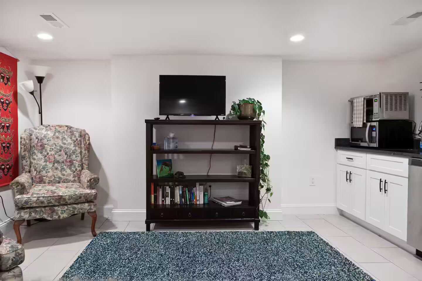 Ein Wohnzimmer mit einem floralen Sessel, einer dunkelholznen Bücherwand mit Flachbildfernseher und einer Küchenzeile inklusive Mikrowelle, sowie einem blauen Teppich mit Muster.