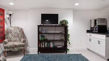 Ein Wohnzimmer mit einem floralen Sessel, einer dunkelholznen Bücherwand mit Flachbildfernseher und einer Küchenzeile inklusive Mikrowelle, sowie einem blauen Teppich mit Muster.