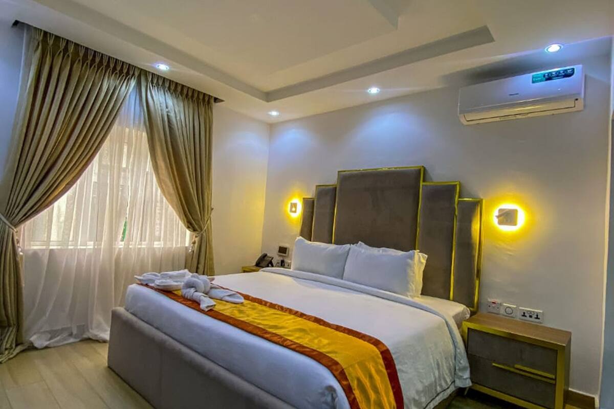 A modern hotel room with a large bed, a bedside table, an air conditioning unit, and a window with curtains.