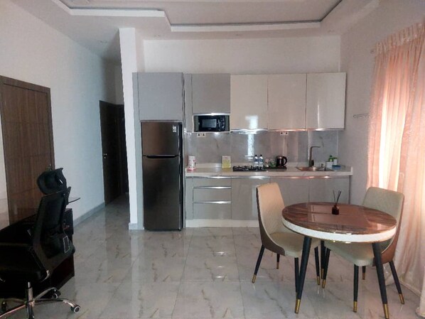 A compact kitchen with a refrigerator, microwave, and stove. There is a dining table with chairs and a small office chair. - Sapphire Hotel and Apartments (Asese)