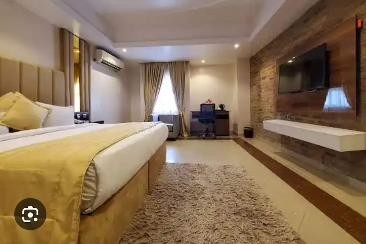 A modern hotel room with a large bed, a desk, a TV mounted on a brick wall, and a large window with curtains.