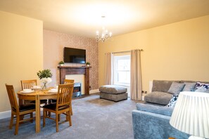 A living room with a fireplace, a dining table with chairs, a sofa, and a television mounted on the wall. - Rooms at the Vault (Buxton)