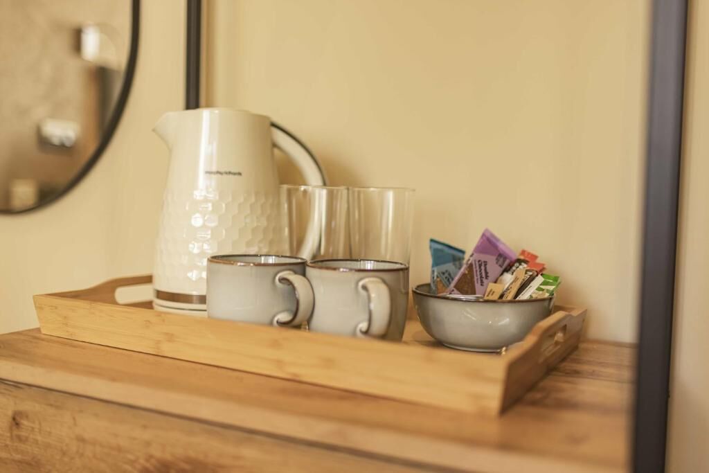 A wooden tray with a white pitcher, two clear glasses, and a bowl containing colorful packets on a wooden surface.