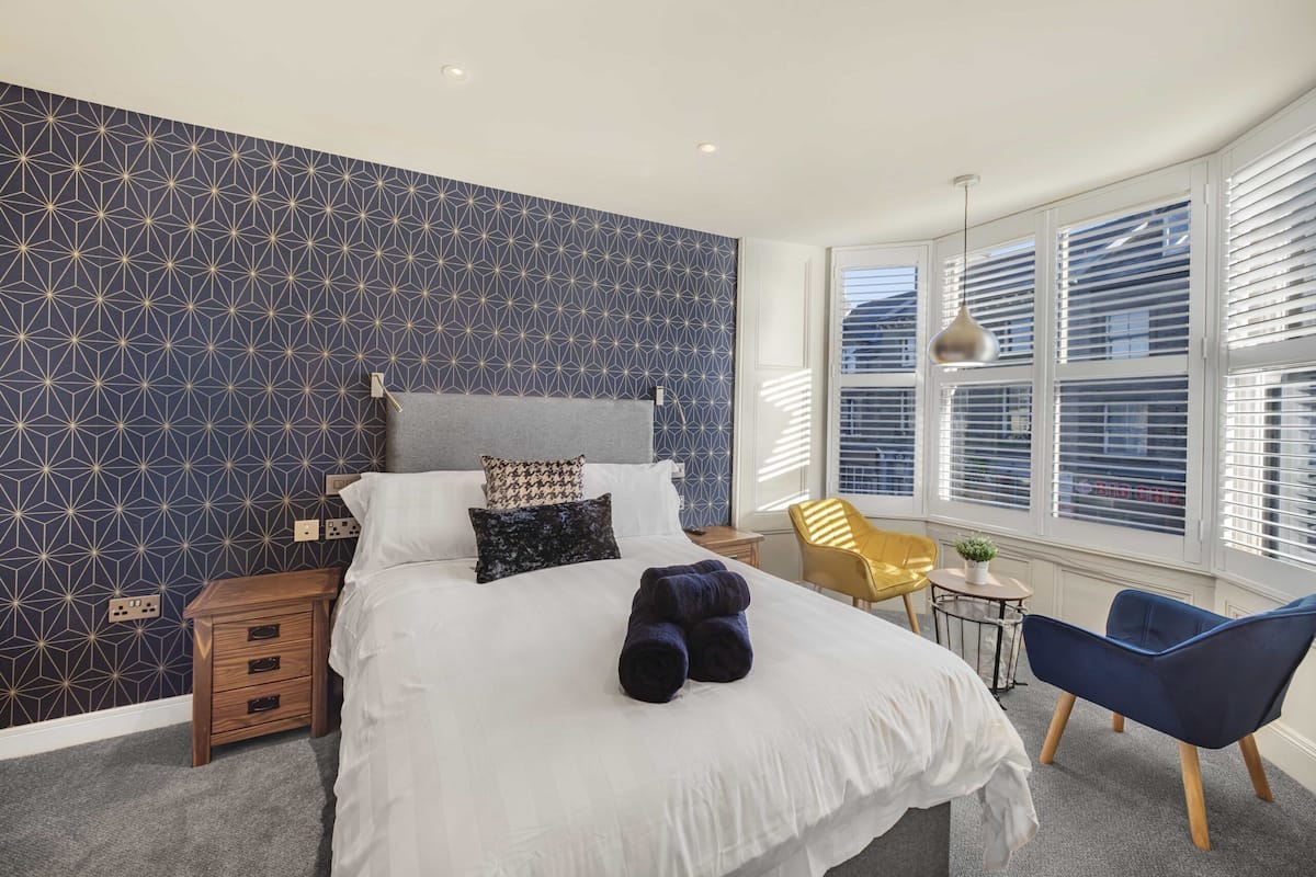 A bedroom with a geometric patterned wall, a bed with white bedding, a bedside table, a chair, and a window with blinds.