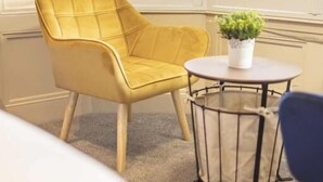 Un sillón amarillo junto a una ventana con persianas, una mesa redonda con una planta en maceta y una pequeña mesa redonda con estructura metálica.