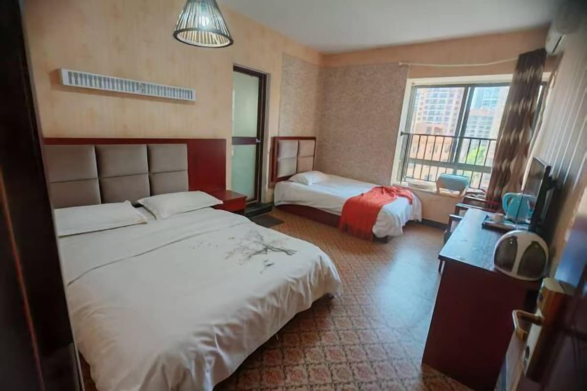 A hotel room with two beds, a desk with a chair, a window with a view of buildings, and a ceiling fan.