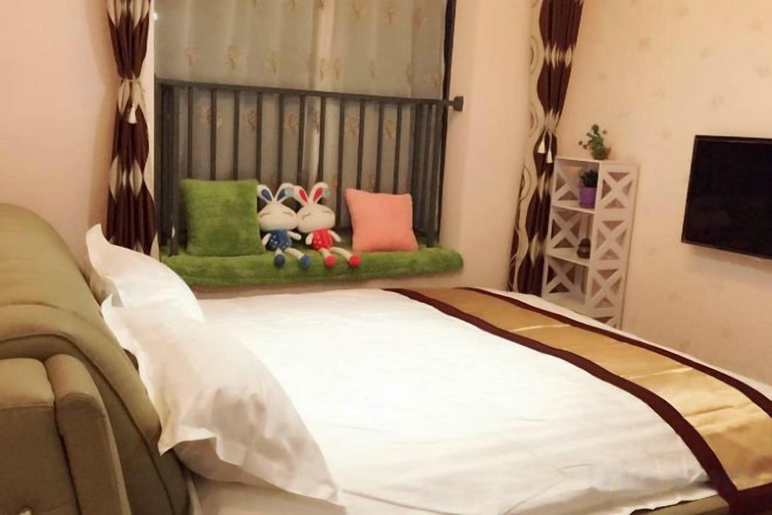 A bedroom with a bed, a sofa, a window with curtains, a small table with a plant, and a TV on the wall.