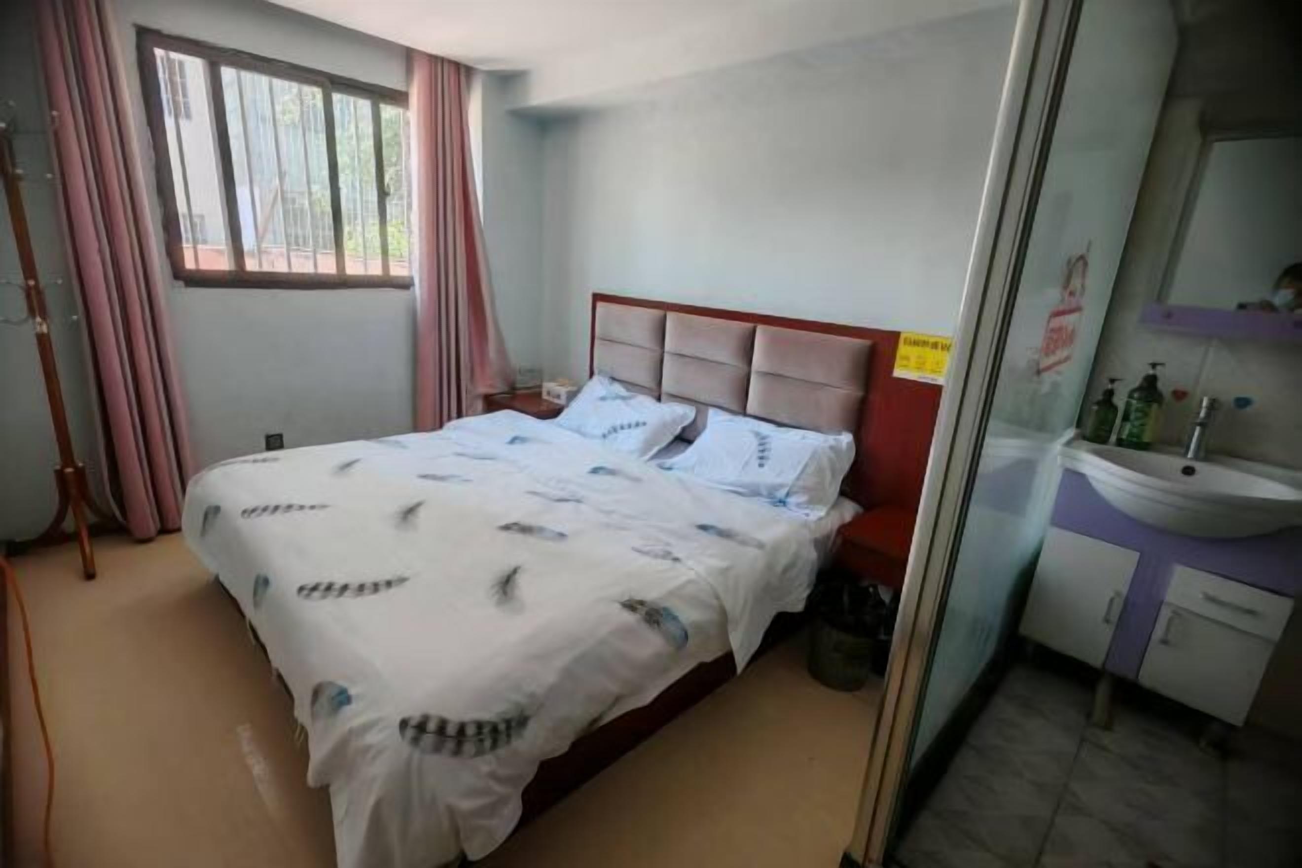 A hotel room with a bed, a bedside table, a window with curtains, a bathroom with a sink and mirror, and a trash bin.