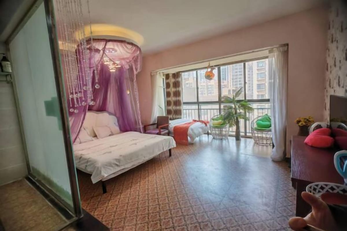 A bedroom with a bed, a window with a view of buildings, a balcony with chairs, and a plant.