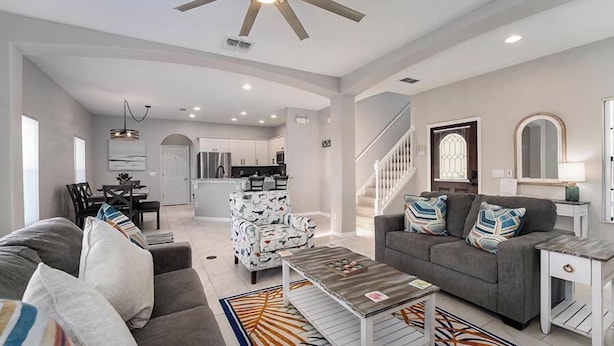 A spacious living room with a gray sofa, a coffee table, and patterned chairs. A staircase leads to a kitchen area with white cabinets. - Poolside Paradise on Sandy Point - with Games Room (Clermont)