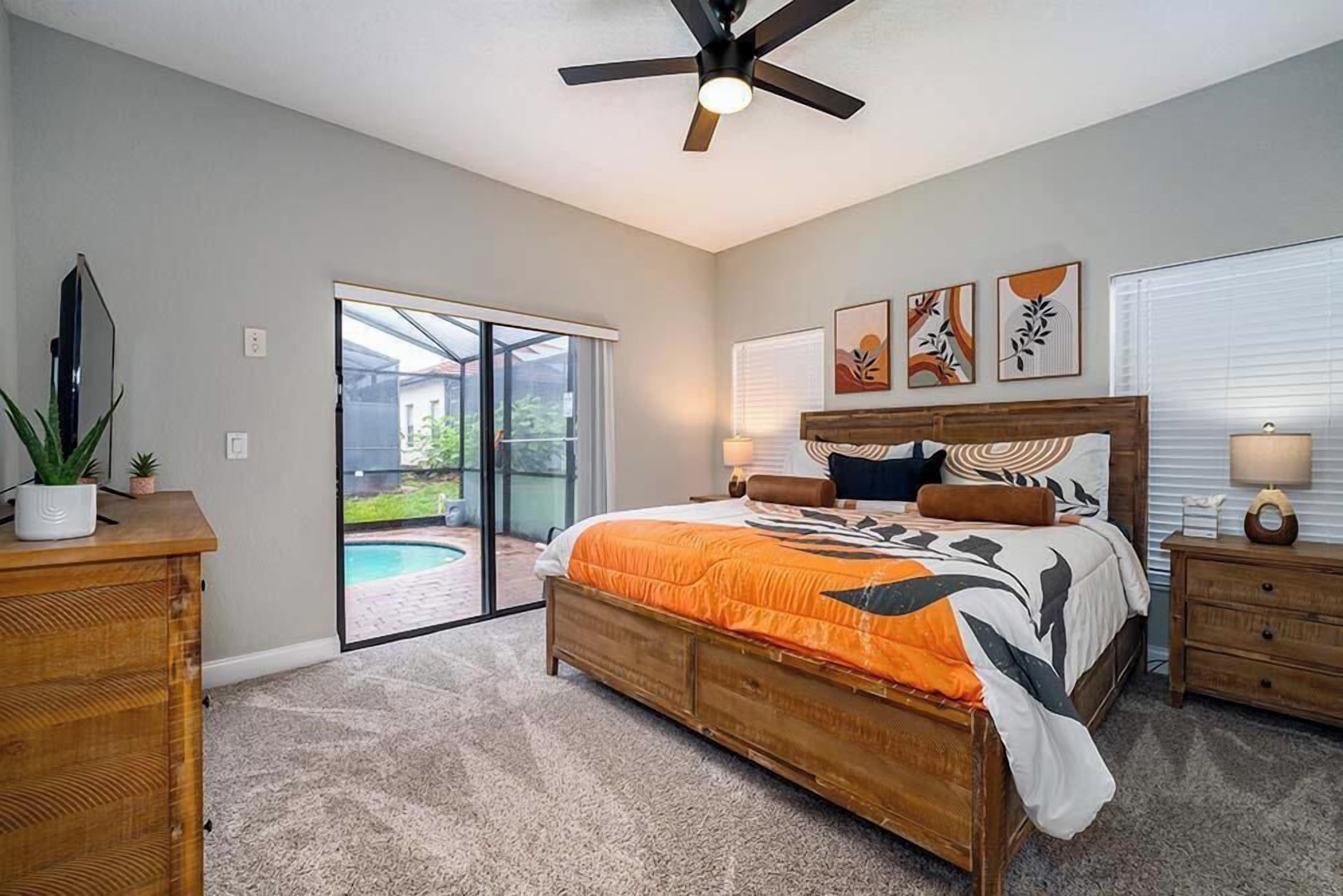 A bedroom with a large bed, a ceiling fan, a TV, a sliding glass door to a patio, and artwork on the wall.