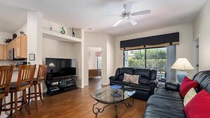 A living room with a black leather sofa, a glass coffee table, wooden chairs, a TV, and a ceiling fan. - Caribbean Breeze Villa - Private Pool & Spa (Clermont)