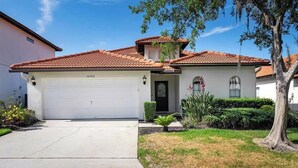 A single-story house with a red-tiled roof, a white garage, and a front yard with a small tree and shrubs. - Caribbean Breeze Villa - Private Pool & Spa (Clermont)