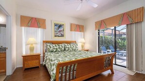 A bedroom with a wooden bed, two bedside tables with lamps, a ceiling fan, and a view of the outdoors through a sliding glass door. - Caribbean Breeze Villa - Private Pool & Spa (Clermont)
