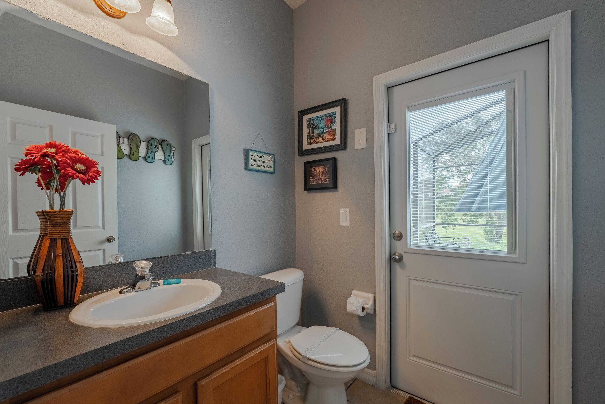 Une salle de bain avec un lavabo blanc, des meubles en bois, un miroir, un vase avec des fleurs, des toilettes et une porte avec une fenêtre.