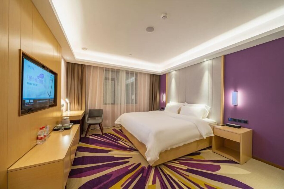 A hotel room with a bed, a desk, a chair, a TV, and a purple accent wall.
