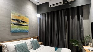 Habitación de hotel con cama, un cuadro en la pared, equipo de aire acondicionado, cortina, planta y escritorio con teléfono.