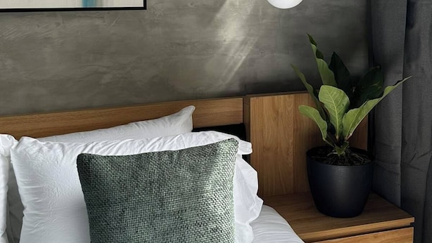 Un dormitorio moderno con una pintura de paisaje urbano, una lámpara colgante, un cabecero de madera, una planta en maceta y una mesita de noche.