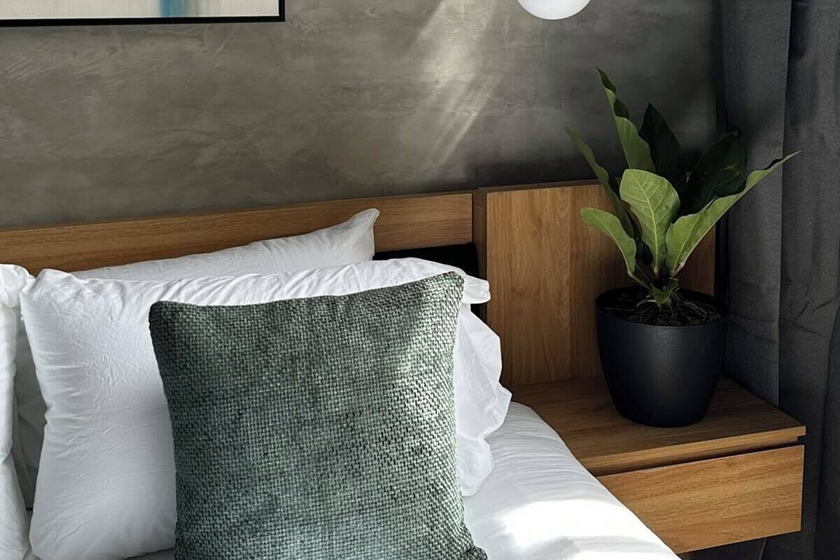 A modern bedroom with a cityscape painting, a hanging light fixture, a wooden headboard, a potted plant, and a bedside table.