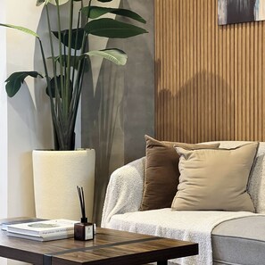 Una sala de estar moderna con un sofá, una planta grande en maceta, una lámpara y un libro sobre una mesita.
