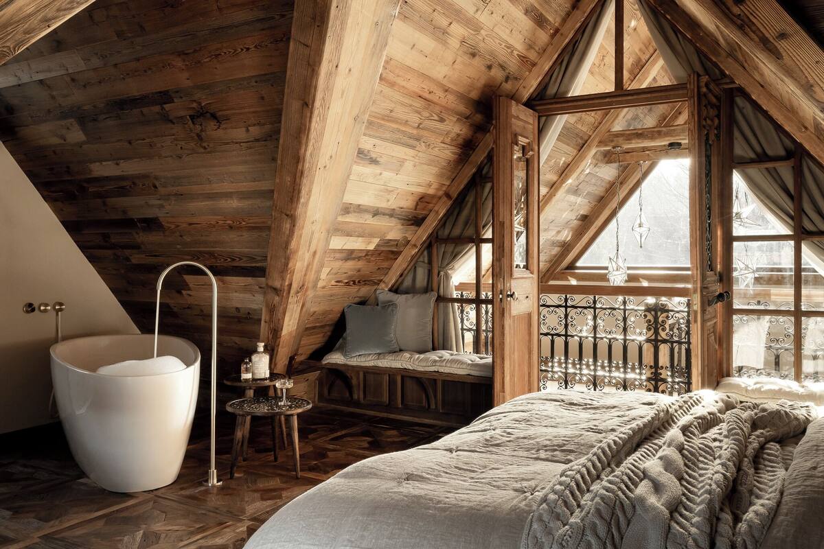 A cozy attic bedroom with a large bathtub, a wooden ceiling, and a window with a view.